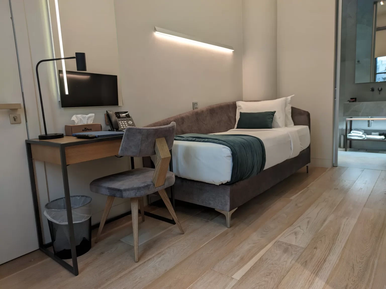 A compact modern Hellenic Vibes Smart hotel room with a single bed featuring white linens and a teal throw, next to a minimalist wooden desk with an upholstered chair, desk lamp, phone, and small mounted TV. Light wood flooring and soft wall lighting create a clean, contemporary look, with an open bathroom area visible in the background Hellenic Vibes Smart hotel, smart hotel Athens, hotel near Syntagma, hotel near Acropolis, hotel near Monastiraki, central Athens accommodation, business room Athens, hotel with workspace Athens, digital nomad Athens