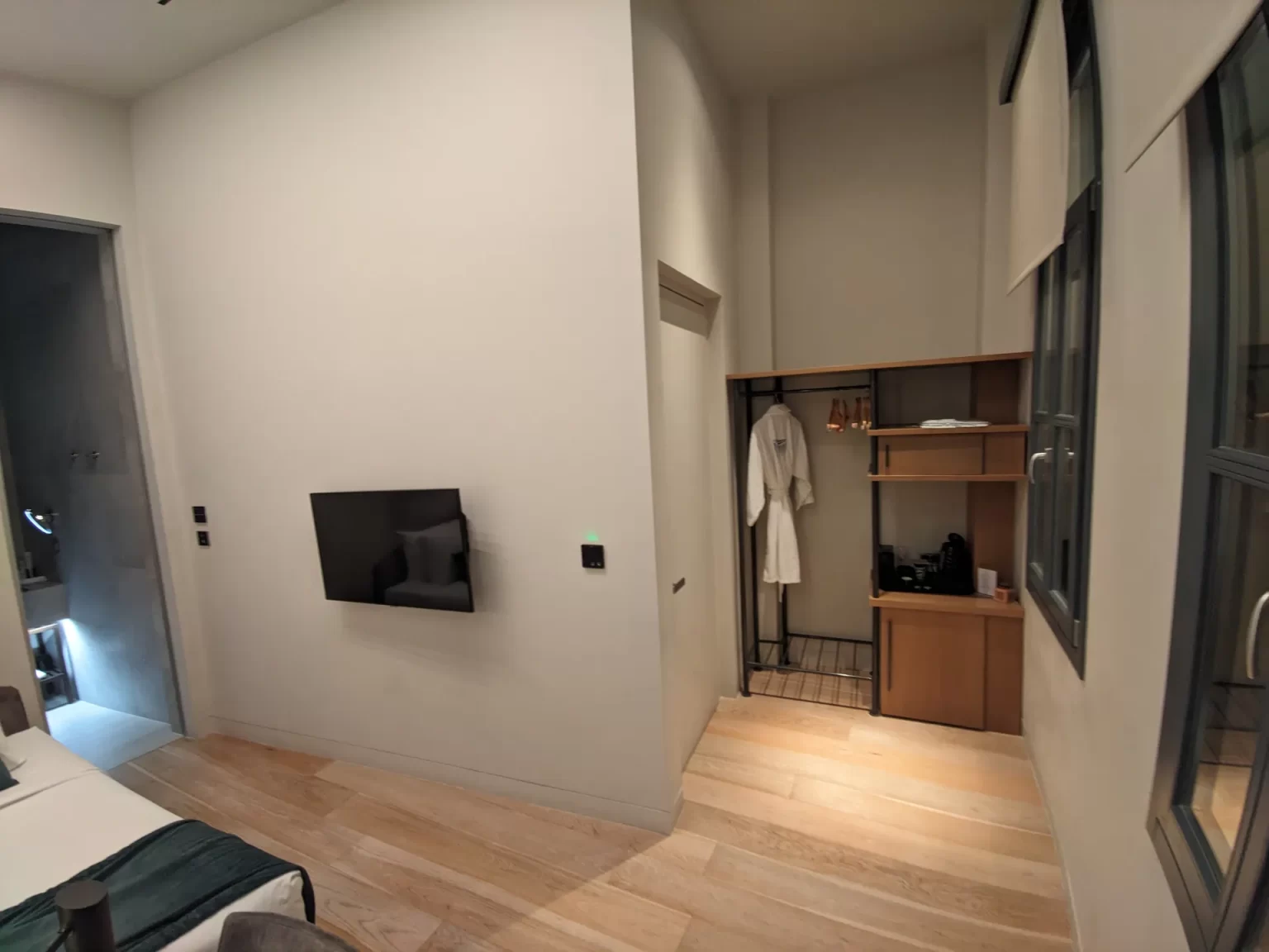 A modern hotel room with light wood flooring, featuring a wall mounted TV, an open wardrobe with shelves, hangers, and a bathrobe, along with a coffee and tea station. Soft neutral walls and a large window create a bright, minimalist atmosphere, with part of the neatly made bed visible in the corner Hellenic Vibes Smart hotel, smart hotel Athens, hotel near Syntagma, hotel near Acropolis, hotel near Monastiraki, central Athens accommodation, business room Athens, hotel with workspace Athens, digital nomad Athens