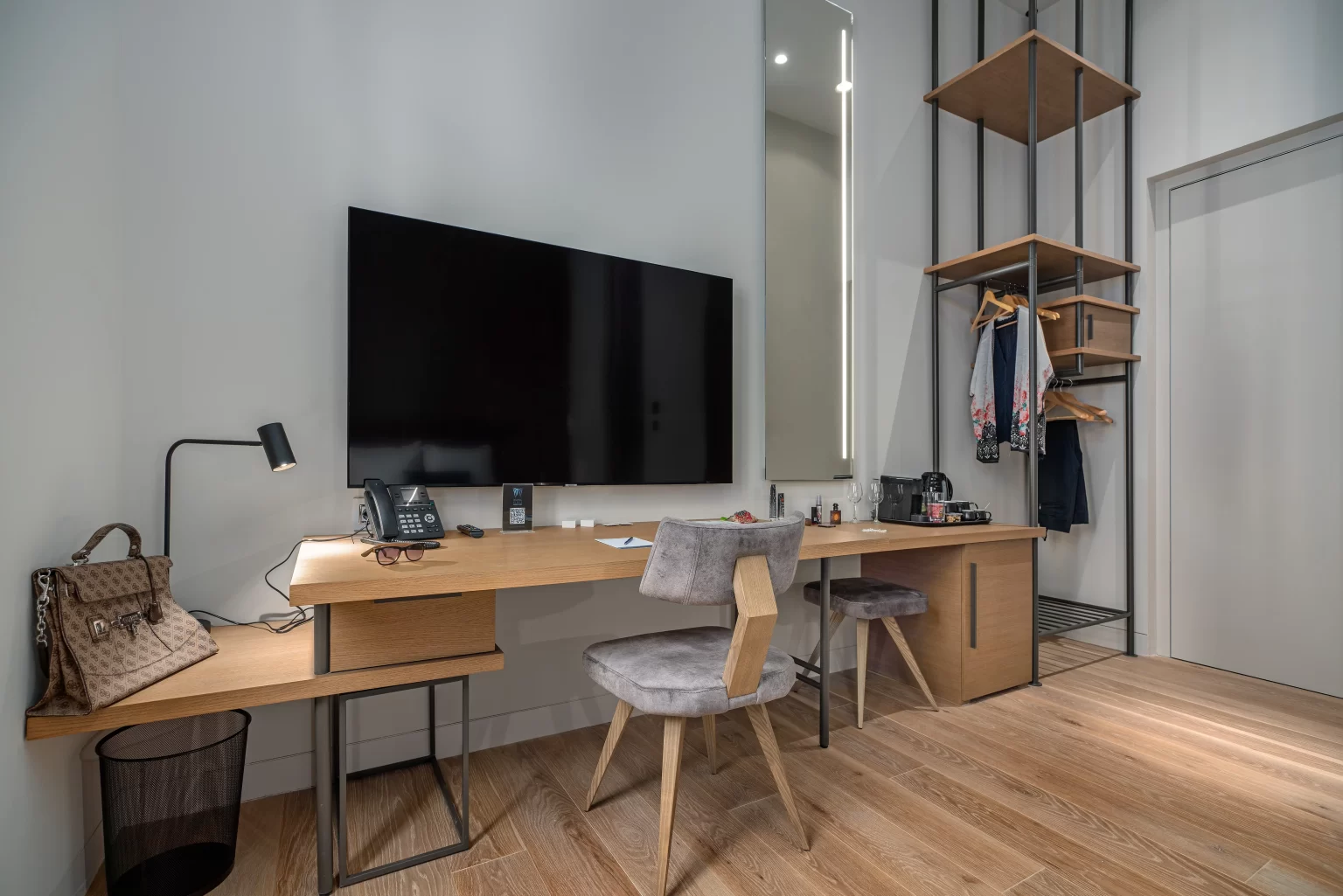 A modern Hellenic Vibes Smart hotel room workspace featuring a long wooden desk with two upholstered chairs, a large wall mounted flat screen TV, a desk lamp, phone, coffee and tea station, and neatly arranged personal items. Open shelving with clothes hanging and warm wood flooring enhance the clean, minimalist design. Hellenic Vibes Smart hotel, smart hotel Athens, hotel near Syntagma, hotel near Acropolis, hotel near Monastiraki, business suite Athens, hotel with workspace Athens, work-friendly suite Athens, digital nomad Athens