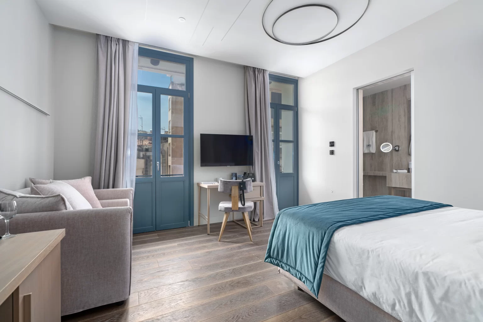 Bright modern Hellenic Vibes Smart hotel room with queen bed, teal throw, sofa, desk, TV, and city-view windows. Hellenic Vibes Smart hotel, smart hotel Athens, hotel near Syntagma, hotel near Acropolis, hotel near Monastiraki, central Athens accommodation,