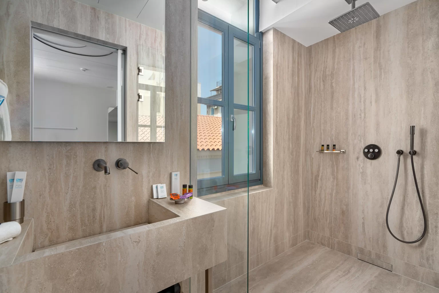 Modern beige marble bathroom in Hellenic Vibes Smart hotel with a sleek sink, large mirror, rainfall shower, and natural light from a window. Hellenic Vibes Smart hotel, smart hotel Athens, hotel near Syntagma, hotel near Acropolis, hotel near Monastiraki, luxury suite bathroom, modern bathroom Athens hotel, walk-in shower Athens, Spa-style suite bathroom, boutique hotel bathroom Athens