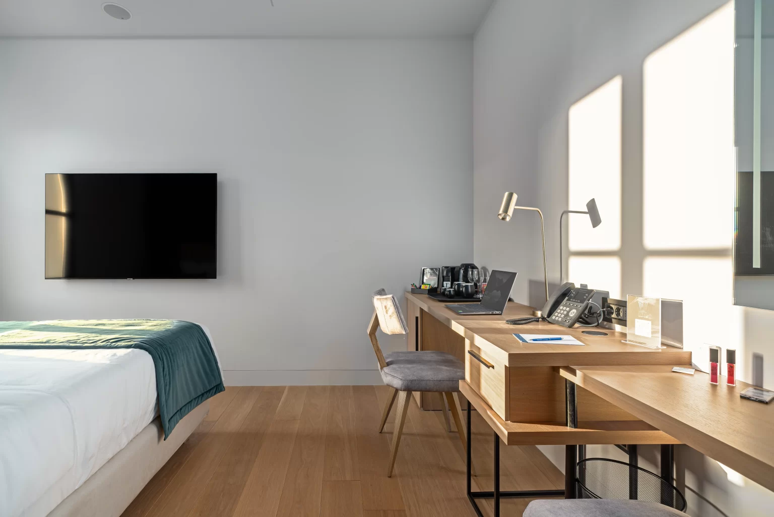 A Hellenic Vibes Smart hotel room workspace with a long wooden desk, ergonomic chair, desk lamp, coffee and tea station, and a laptop set up beside a wall-mounted flat screen TV and neatly made bed with a teal blanket. Sunlight casts geometric shadows on the white wall, highlighting the room’s clean, minimalist design. Hellenic Vibes Smart hotel, smart hotel Athens, hotel near Syntagma, hotel near Acropolis, hotel near Monastiraki, Athens city center suites, business suite Athens, hotel with workspace Athens, work-friendly hotel Athens, digital nomad Athens