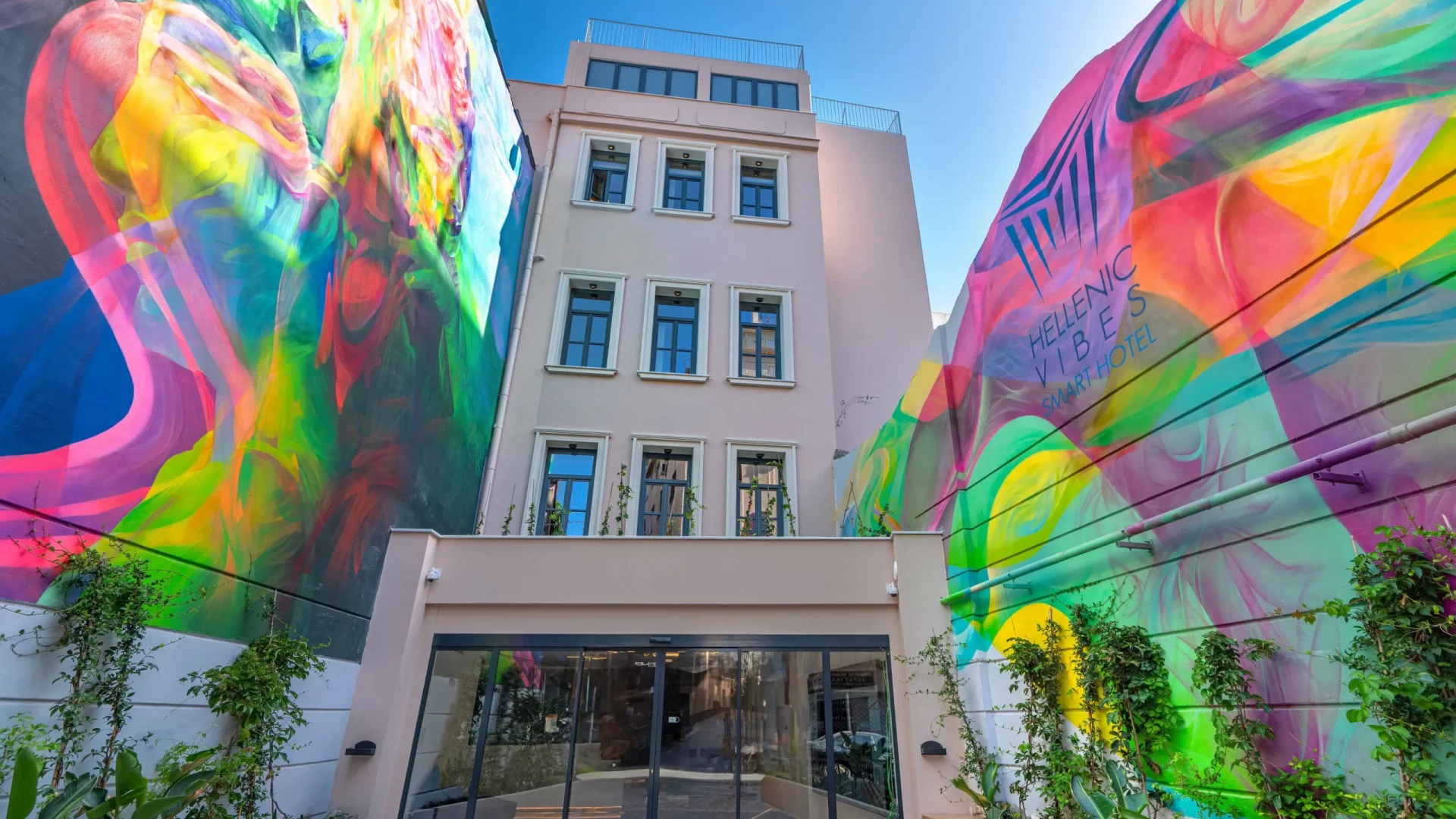 Vibrant exterior of the Hellenic Vibes Smart hotel featuring a modern façade framed by large, colorful street art murals and lush greenery at the entrance, creating a striking and contemporary welcome for guests. Hellenic Vibes Smart hotel, hotel near Syntagma, hotel near Acropolis, hotel near Moanstiraki, smart hotel Athens, central Athens accommodation, Athens city center attractions, walkable Athens hotel location